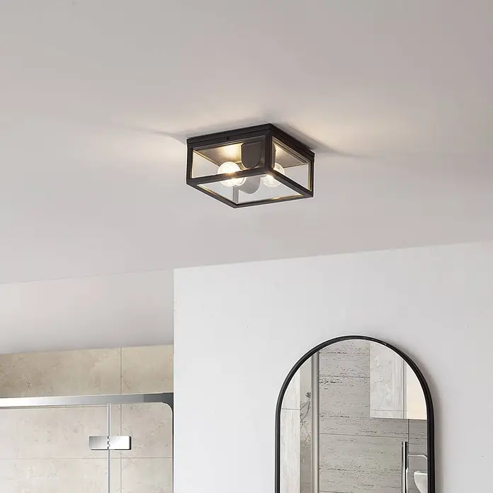 Lindby Außenwand- & Deckenleuchte Kremena - schwarz, klar / transparent, IP44 - Quadratische Deckenleuchte aus schwarzem Metall mit klaren Glaspaneelen und zwei beleuchteten Glühbirnen, in einem modernen Badezimmer installiert.