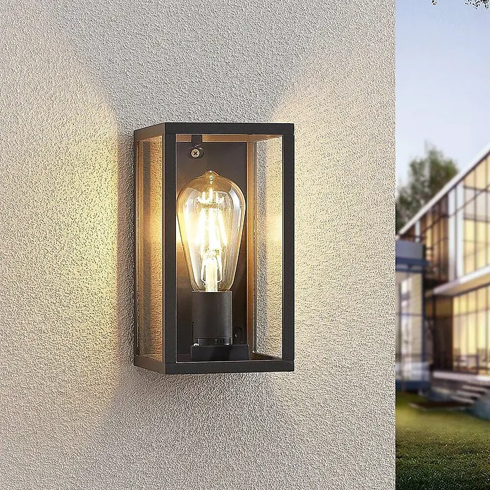Lindby Außenwandleuchte Filimon - 12 x 12 x 22 cm, schwarz, schwarz, IP44 - Schwarze Außenwandleuchte mit Vintage-Glühbirne, beleuchtet eine strukturierte Wand neben einem modernen Haus.