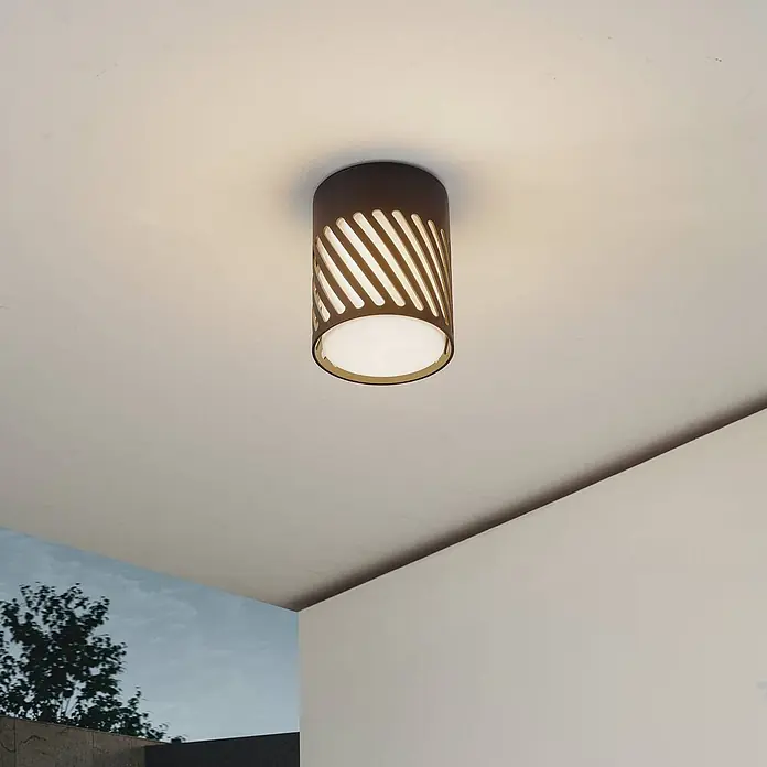 Lindby Außenwand- & Deckenleuchte Lurinda - schwarz, IP44 - Dunkelbraune zylindrische Deckenleuchte mit diagonalen Aussparungen, an einer Decke montiert, spendet nach unten und seitlich gemustertes Licht.