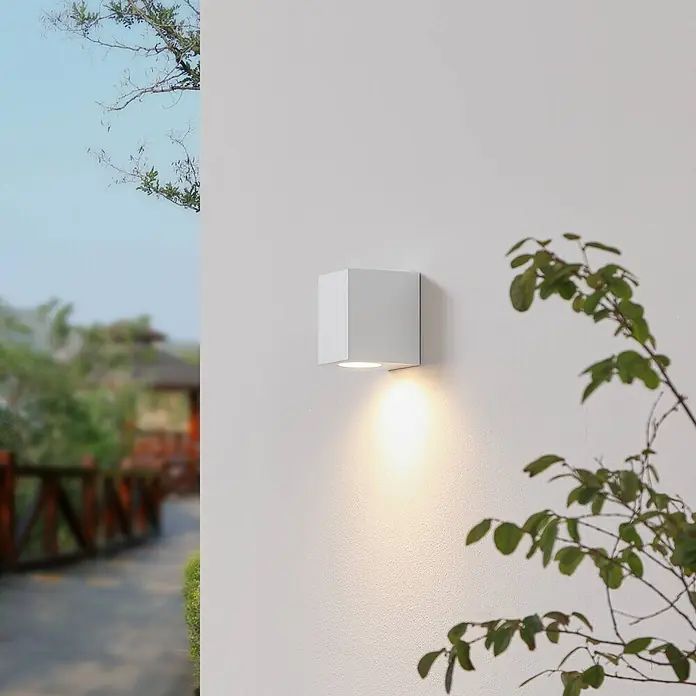 Prios Außenwandleuchte Irfan - weiß / opal, IP44 - Weiße, quaderförmige Außenwandleuchte beleuchtet eine strukturierte Wand mit warmem Licht, in einer Außenszene mit Blättern und einem unscharfen Weg.