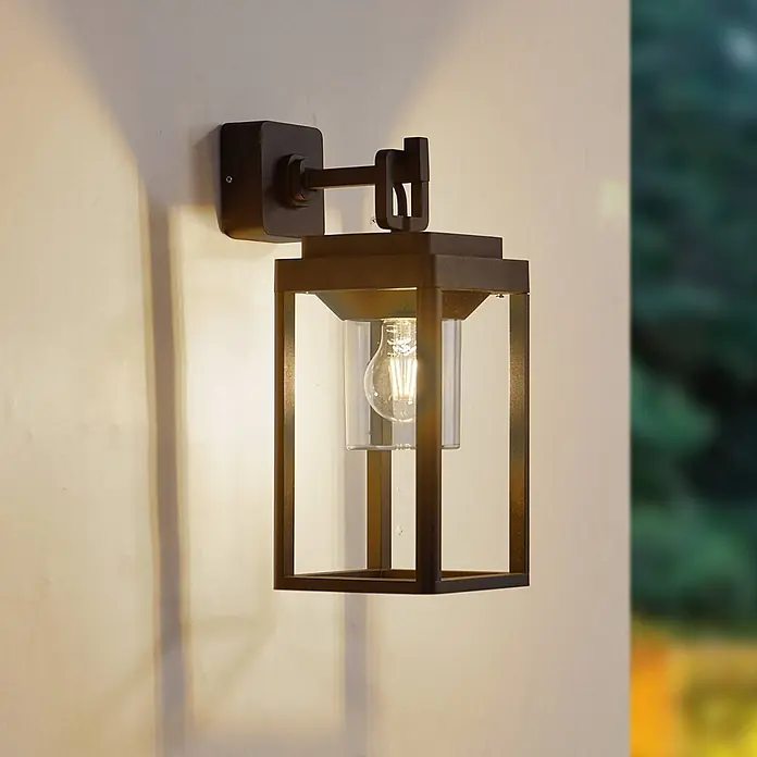 Lucande Außenwandleuchte Milanka - schwarz, schwarz, IP65 - Dunkle Metallrahmen-Außenwandleuchte mit klarem Glaszylinder und sichtbarer Glühbirne.