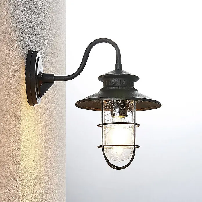 Lindby Außenwandleuchte Kyan - schwarz, IP44 - Schwarze Metall-Außenwandleuchte mit klarem, blasigem Glas, montiert und beleuchtet, wirft warmes Licht auf eine strukturierte Wand.