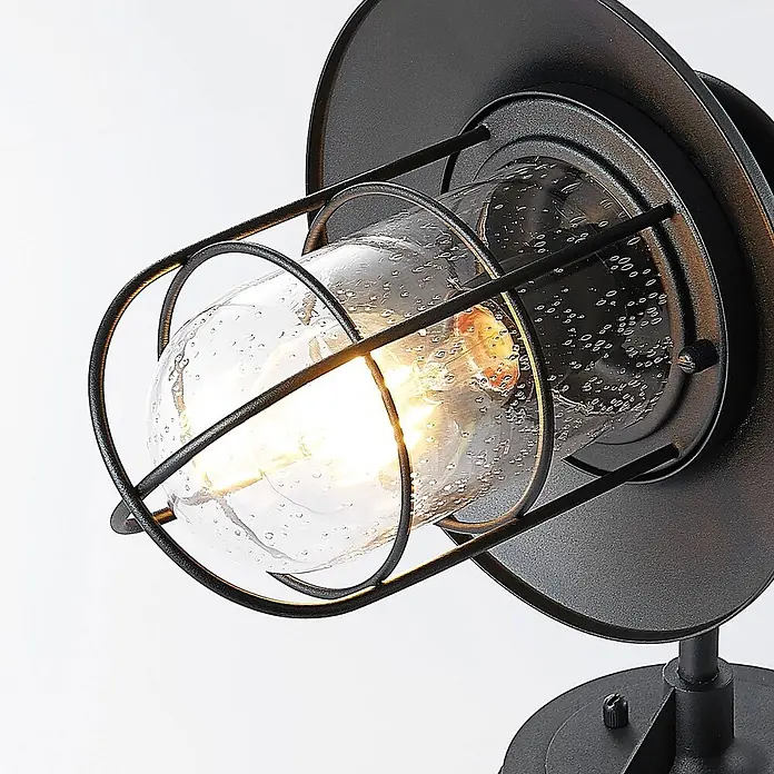 Lindby Außenwandleuchte Kyan - schwarz, IP44 - Nahaufnahme einer schwarzen Metall-Wandleuchte im Industriedesign mit Samenglas und warm leuchtender Glühbirne.