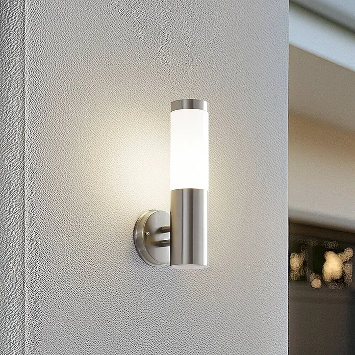 Lindby Außenwandleuchte Kristof - alu / grau / zink, alu / grau / zink, IP44 - Zylindrische Wandleuchte aus gebürstetem Edelstahl mit mattweißem Diffusor, beleuchtet und an einer strukturierten Wand montiert.