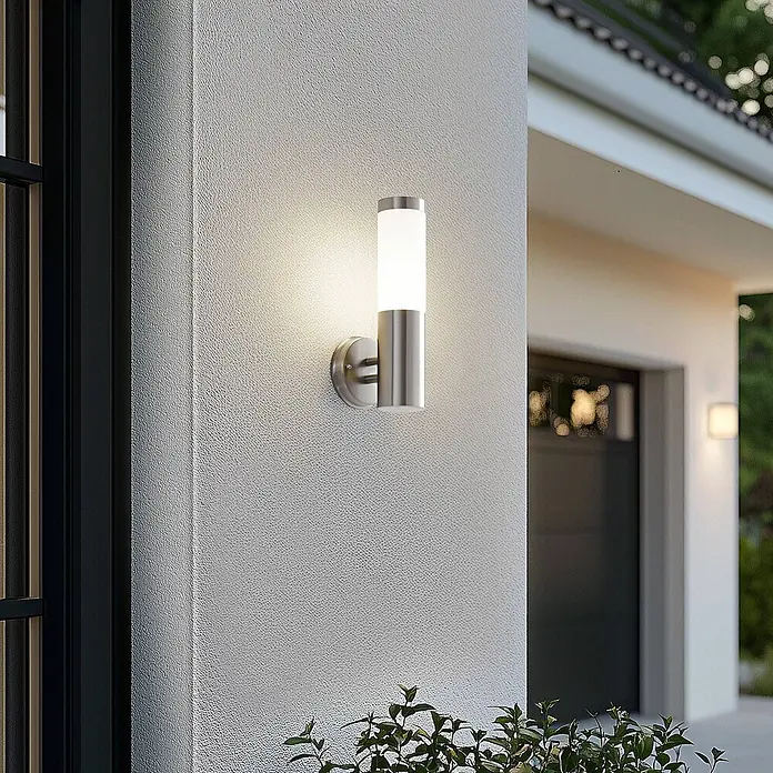 Lindby Außenwandleuchte Kristof - alu / grau / zink, alu / grau / zink, IP44 - Außenwandleuchte aus gebürstetem Edelstahl mit zylindrischem weißem Diffusor, beleuchtet an einer Außenwand.