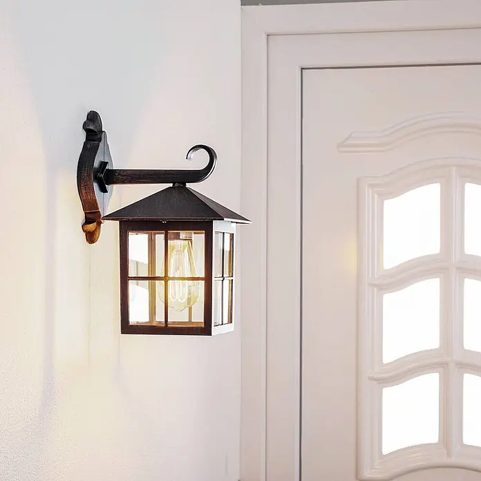 Lindby Außenwandleuchte Ida - braun / rost, braun / rost, IP23 - Beleuchtete Wandlaterne aus dunklem Metall und Glas an heller Wand neben weißer Tür.