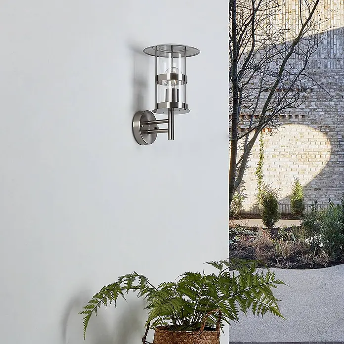 Lindby Außenwandleuchte Noemi - alu / grau / zink, alu / grau / zink, IP44 - Außenwandleuchte aus gebürstetem Edelstahl mit Klarglaszylinder, die eine Ziegelwand und einen Garten im Abendlicht beleuchtet.