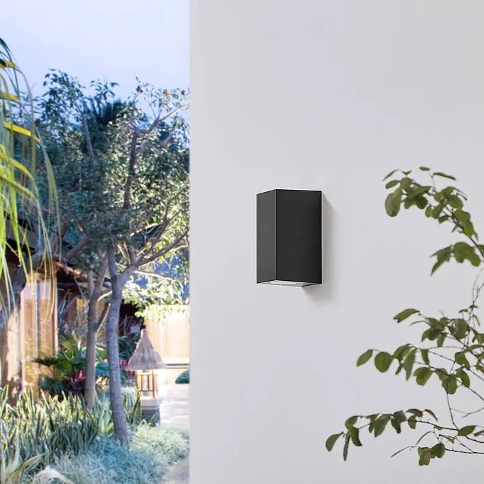 Prios Außenwandleuchte Irfan - schwarz, IP44 - Schwarze rechteckige Außenwandleuchte an einer weißen Wand, mit Gartenblick im Hintergrund.