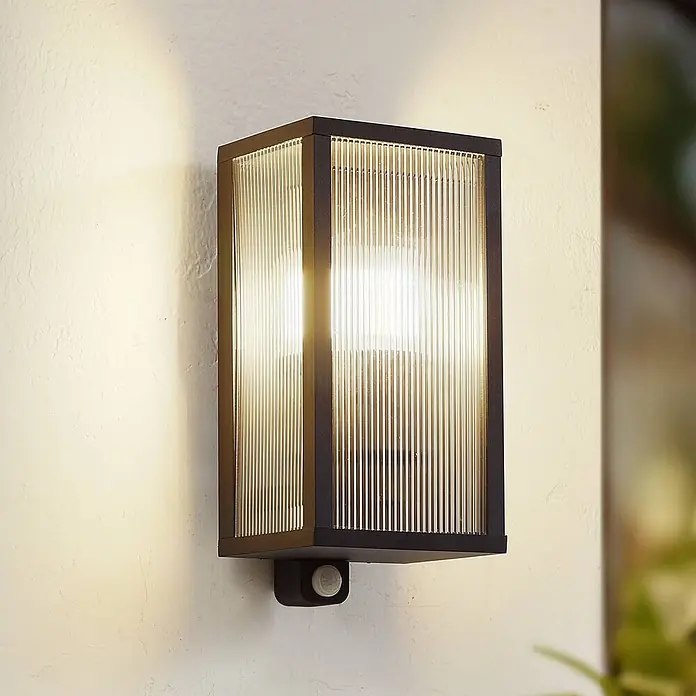 Lindby Außenwandleuchte Peldar - 13,5 x 14 x 29 cm, schwarz, IP44 - Schwarze Außenwandleuchte mit gerippten Glaspaneelen und integriertem Bewegungsmelder, beleuchtet an einer Wand.