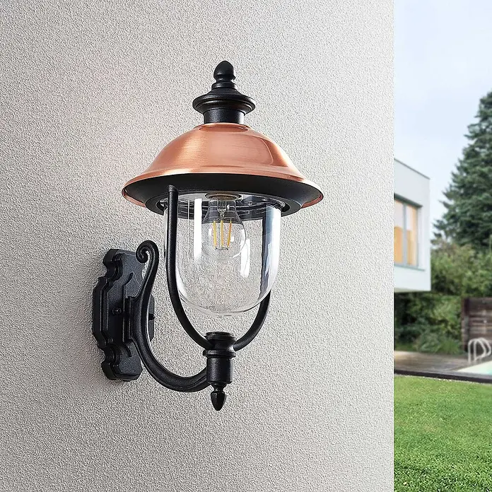 Lindby Außenwandleuchte Clint - 30 x 24,9 x 43,5 cm, kupfer, IP44 - Außenwandleuchte mit Kupferschirm, schwarzem Metallrahmen und klarem Glasdiffusor, an einer strukturierten Wand montiert.