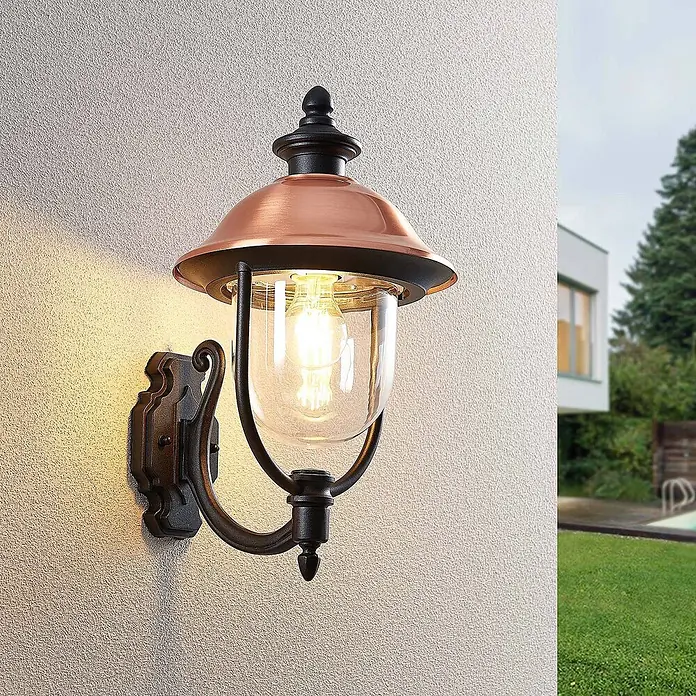 Lindby Außenwandleuchte Clint - 30 x 24,9 x 43,5 cm, kupfer, IP44 - Kupferfarbene Außenwandleuchte mit schwarzem Gestell, die an einer strukturierten Wand warm leuchtet.
