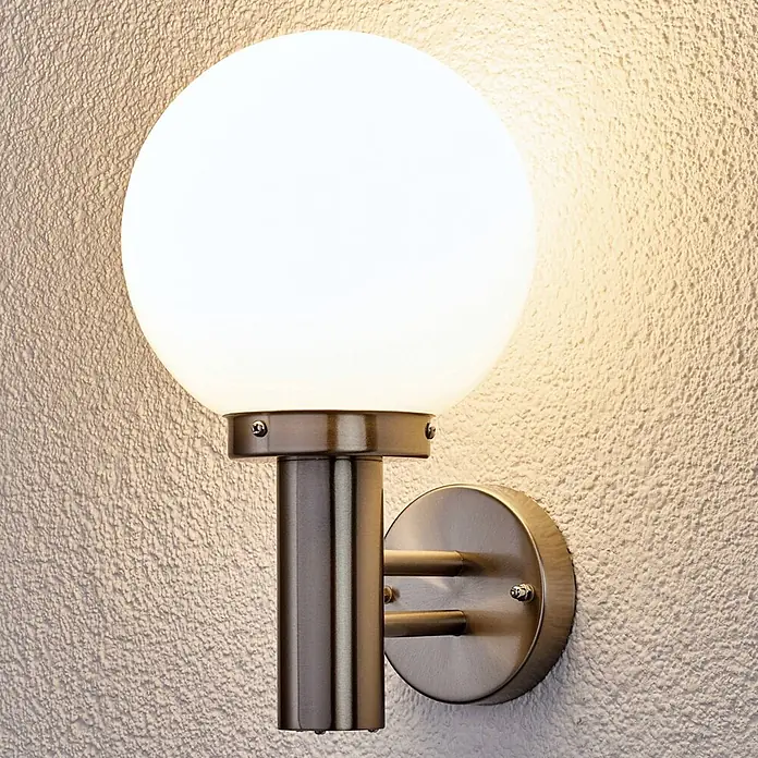 Lindby Außenwandleuchte Nada - alu / grau / zink, IP44 - Wandmontierte Außenleuchte mit kugelförmigem weißem Diffusor und bronzefarbenem Metallsockel, die eine strukturierte Wand beleuchtet.