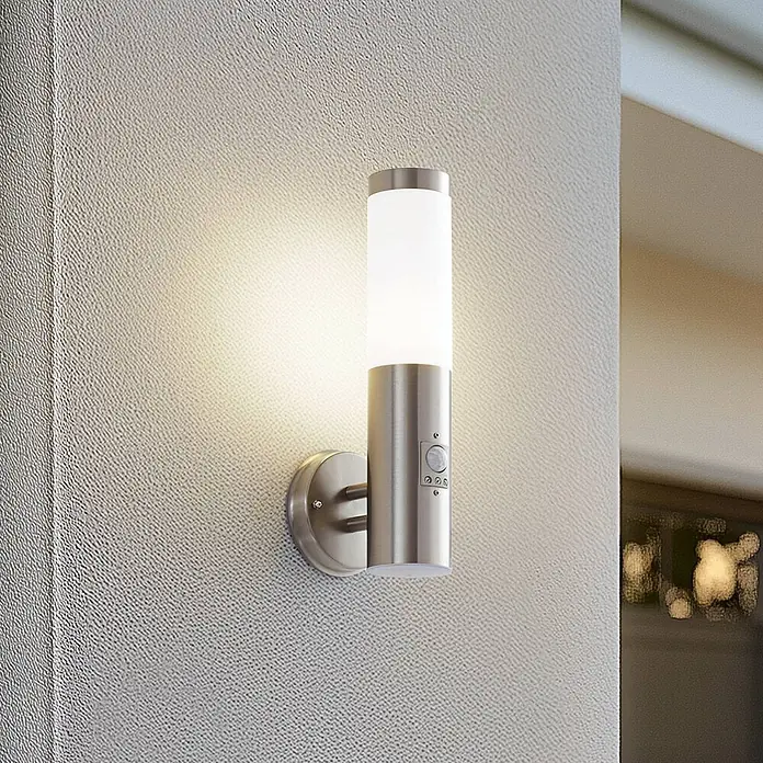 Lindby Außenwandleuchte Kristof - alu / grau / zink, alu / grau / zink, IP44 - Beleuchtete Wandleuchte aus gebürstetem Edelstahl mit weißem Diffusor und Bewegungssensor, an einer strukturierten Wand montiert.