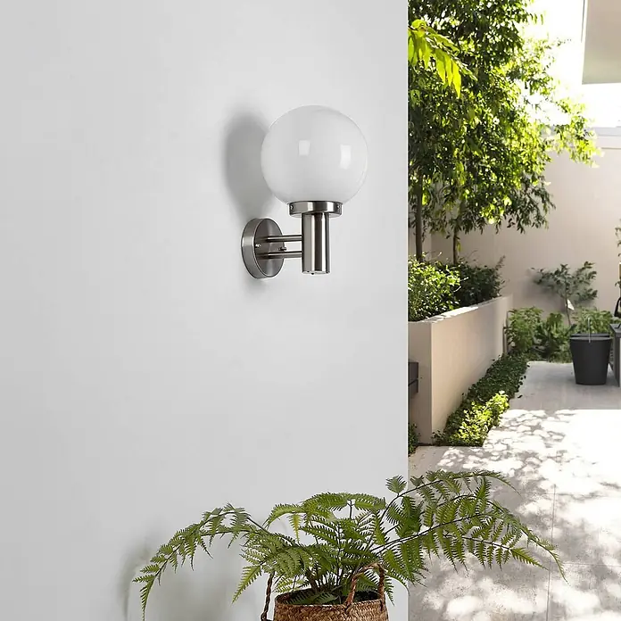 Lindby Außenwandleuchte Nada - alu / grau / zink, IP44 - Wandleuchte aus gebürstetem Metall mit kugelförmigem, weißem Glasschirm, platziert in einem Garten.