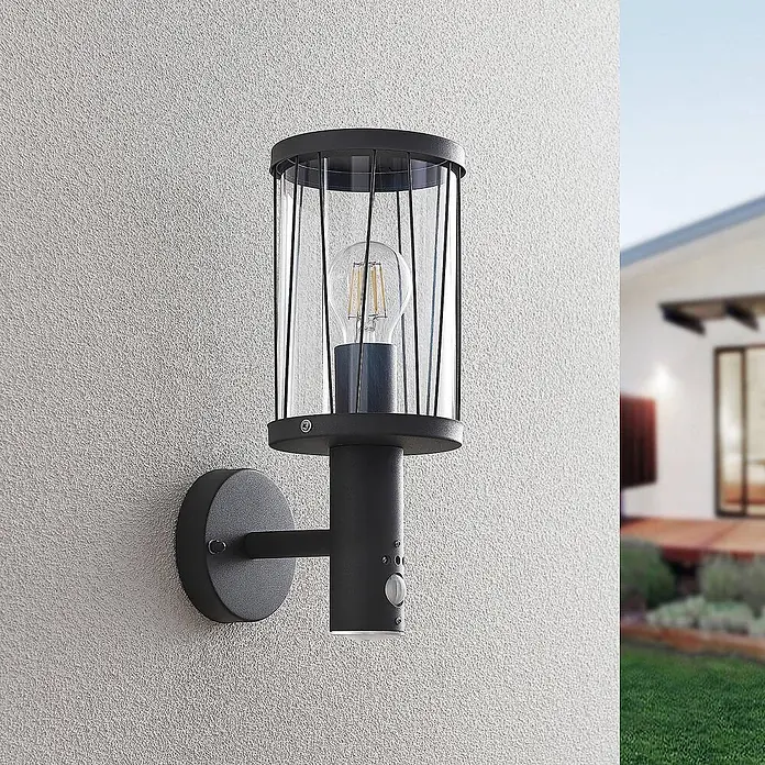 Lindby Außenwandleuchte Yonan - schwarz, schwarz, IP44 - Schwarze Außenwandleuchte mit zylindrischem Glas- und Metallkäfig, sichtbarer Glühlampe und Bewegungssensor, an strukturierter Hauswand montiert.