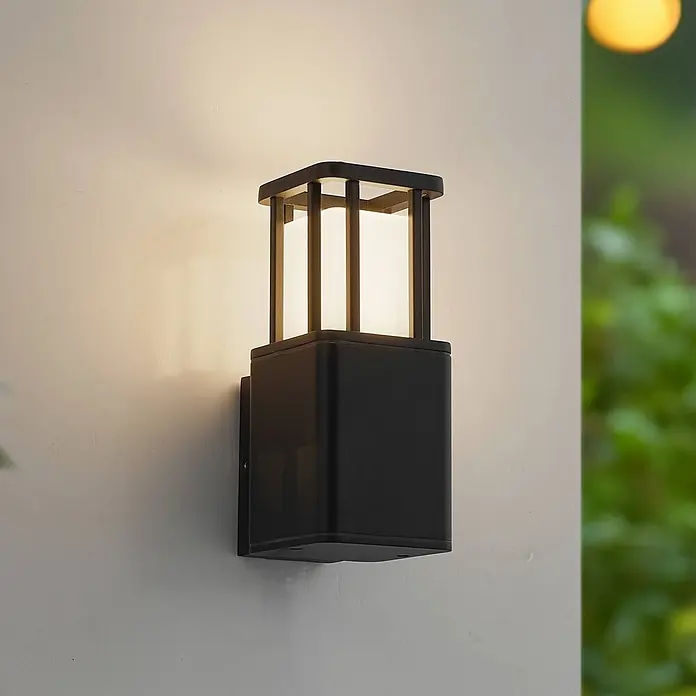Lucande Außenwandleuchte Dakil - schwarz, schwarz, IP65 - Schwarze Außenwandleuchte mit quadratischem, käfigartigem Aufsatz, die warmes Licht auf eine helle Wand wirft.