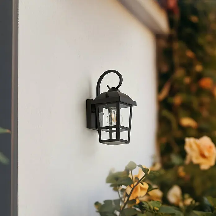 Lindby Außenwandleuchte Eloane - 16 x 13 x 29 cm, schwarz, schwarz, IP44 - Schwarze Außenwandleuchte mit Klarglas und LED-Glühbirne, an einer hellen Wand inmitten von Grün montiert.