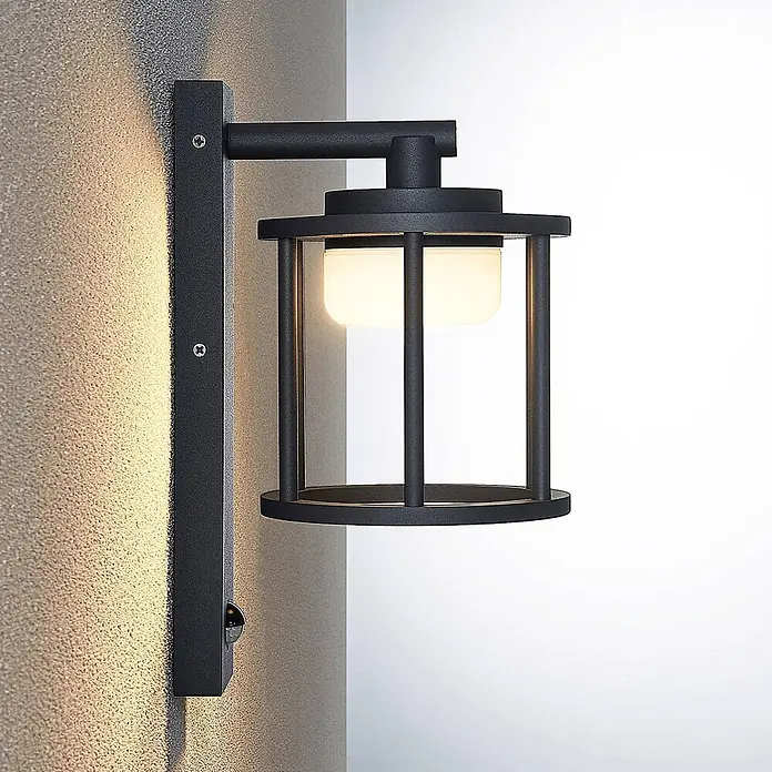 Lucande Außenwandleuchte Berenike - alu / grau / zink, alu / grau / zink, IP44 - Schwarze Metall-Wandleuchte im Laternenstil mit mattem Diffusor und Bewegungssensor an einer strukturierten Wand.