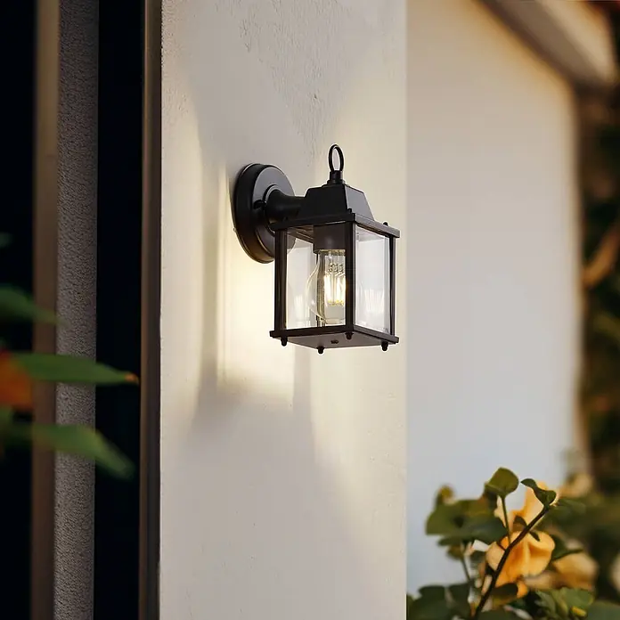 Lindby Außenwandleuchte Rurik - schwarz, IP44 - Schwarze Außenwandleuchte mit Klarglas und freiliegender Glühbirne, an einer hellen Wand montiert.