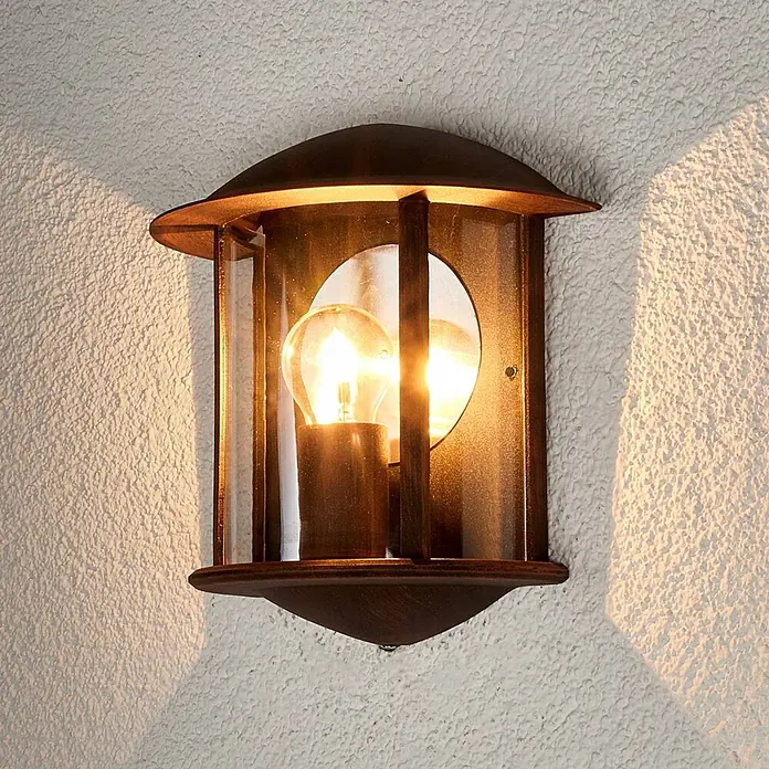 Lindby Außenwandleuchte Maelis - braun / rost, IP44 - Rustikale braune Außenwandleuchte mit Glaspaneelen und einer beleuchteten Glühbirne an einer strukturierten Wand.