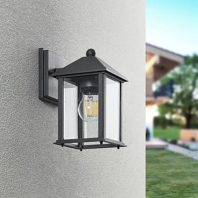 Lindby Außenwandleuchte Giavanna - 18,2 x 14 x 23,3 cm, schwarz, schwarz, IP43 - Schwarze Außenwandleuchte mit klaren Glaspaneelen und sichtbarer LED-Glühbirne, an einer strukturierten Wand montiert.