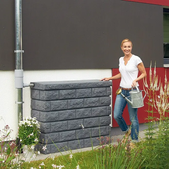 Garantia Exklusive Line Regenspeicher Rocky - 400 l, Darkgranite - Eine Frau steht neben einem grauen, quaderförmigen Wasserbehälter mit Steinmuster und hält eine Gießkanne.