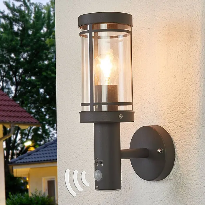 Lindby Außenwandleuchte Djori - alu / grau / zink, IP44 - Anthrazitfarbene Außenwandleuchte mit klarem Glaszylinder und Bewegungsmelder, leuchtend an einer Hauswand montiert.