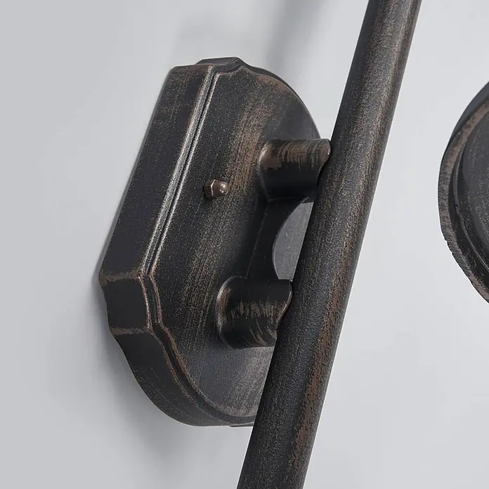 Lindby Außenwandleuchte Daphne - schwarz, schwarz, IP44 - Nahaufnahme eines Wandarmaturteils mit gealterter Bronze-Oberfläche, zeigt eine dekorative Platte und einen Verbindungsarm.
