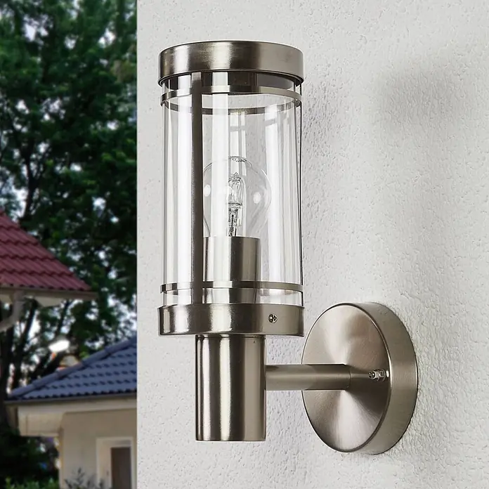 Lindby Außenwandleuchte Djori - alu / grau / zink, IP44 - Außenwandleuchte aus gebürstetem Edelstahl mit zylindrischem Klarglasschirm an einer strukturierten Wand montiert.