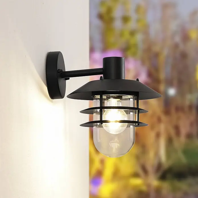 Lindby Außenwandleuchte Meliora - 25,4 x 21,6 x 26,2 cm, schwarz, klar / transparent, IP44 - Schwarze Außenwandleuchte mit Klarglasschirm und sichtbarer Glühbirne, an weißer Wand montiert.