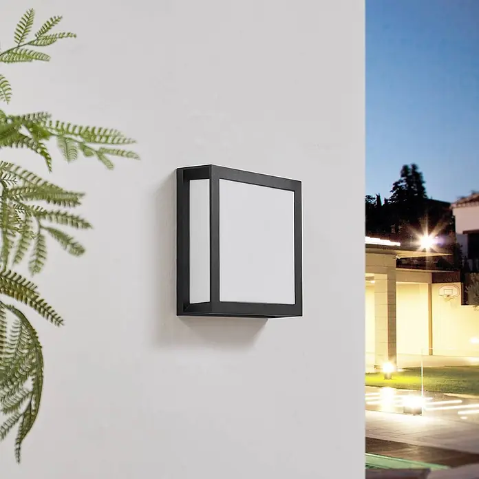Lindby Außenwandleuchte Sivana - schwarz, schwarz, IP44 - Schwarze quadratische Außenwandleuchte mit weißem Diffusor, an einer Wand montiert, beleuchtet eine abendliche Außenszene.