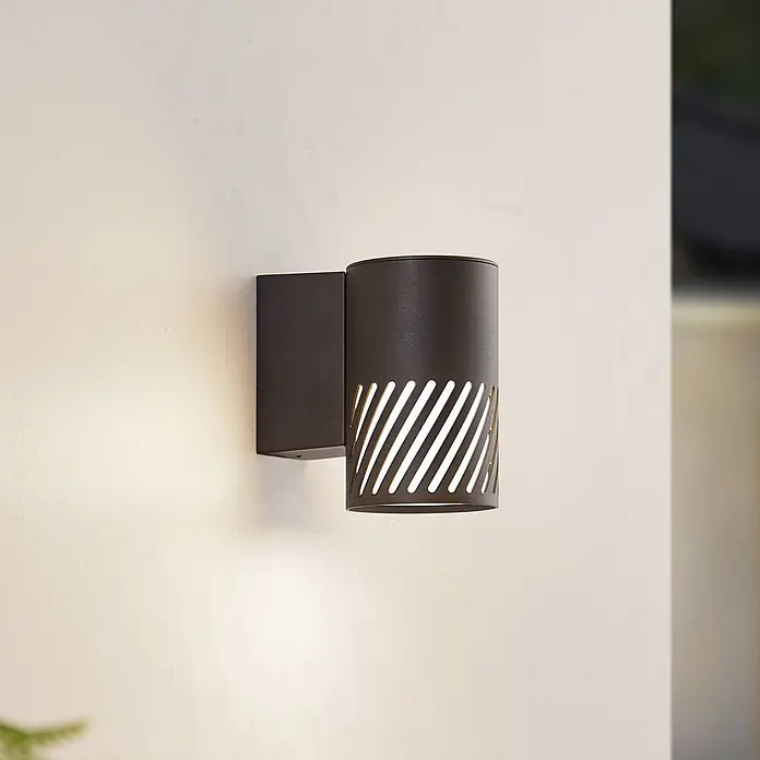 Lindby Außenwandleuchte Lurinda - 16,5 x 10,5 x 16,8 cm, schwarz, schwarz, IP44 - Dunkelgraue zylindrische Außenwandleuchte mit diagonalen Aussparungen, beleuchtet und an einer Wand montiert.