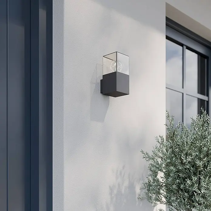 Lucande Außenwandleuchte Keke - schwarz, schwarz, IP44 - Dunkelgraue rechteckige Außenwandleuchte mit Klarglas und dekorativer Glühbirne, an heller Wand neben Fenster und Pflanze montiert.