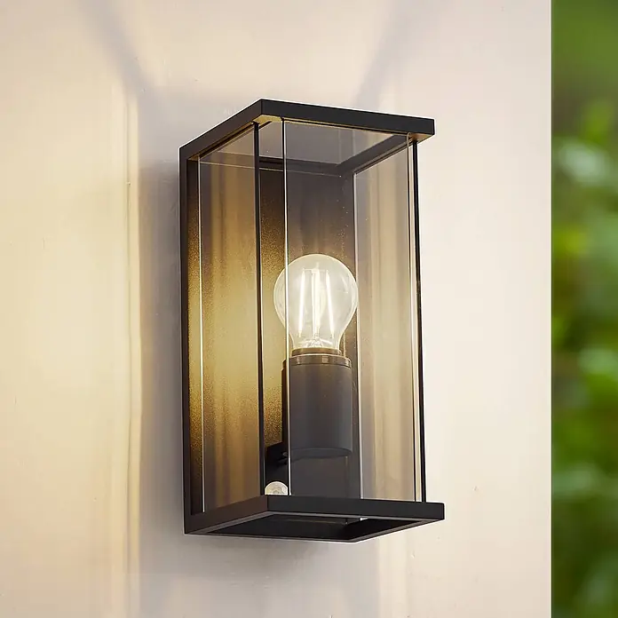 Lucande Außenwandleuchte Annalea - schwarz, IP54 - Schwarze Außenwandleuchte mit Klarglas, beleuchteter LED-Birne und Bewegungssensor, an einer Wand montiert.