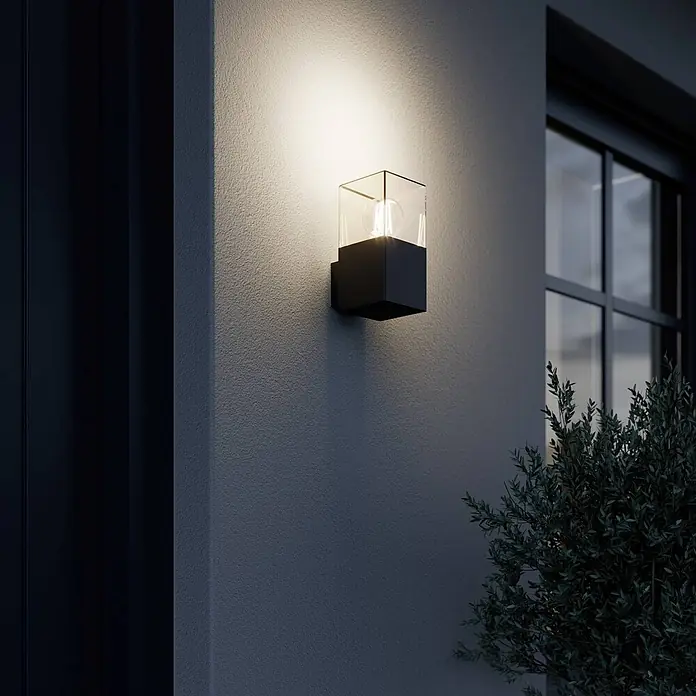 Lucande Außenwandleuchte Keke - schwarz, schwarz, IP44 - Schwarze Außenwandleuchte mit klarem Glasschirm, die eine texturierte Wand bei Nacht beleuchtet.