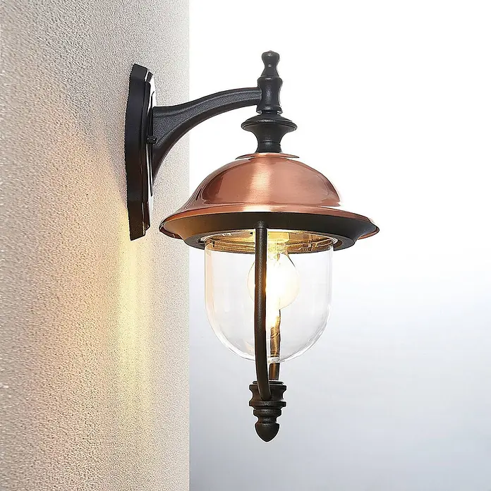 Lindby Außenwandleuchte Clint - kupfer, IP44 - Schwarze und kupferfarbene Wandleuchte mit Klarglas, leuchtend, an einer strukturierten Wand montiert.