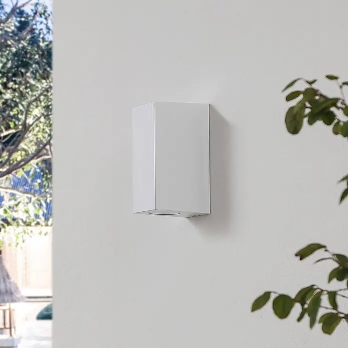 Prios Außenwandleuchte Irfan - 8,9 x 6,8 x 15,5 cm, weiß / opal, IP44 - Weißes rechteckiges Außenwandlicht an einer strukturierten Wand mit verschwommenem Grün.