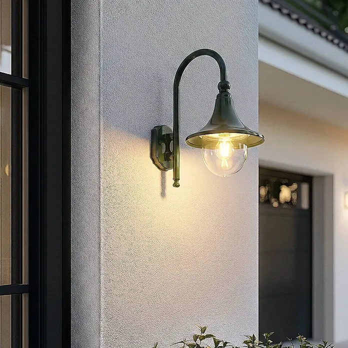Lindby Außenwandleuchte Daphne - 35,5 x 24,5 x 44,2 cm, grün, grün, IP44 - Eine dunkelgrüne Metall-Außenwandleuchte mit klarem Glas und warmer Glühbirne beleuchtet eine strukturierte weiße Außenwand.