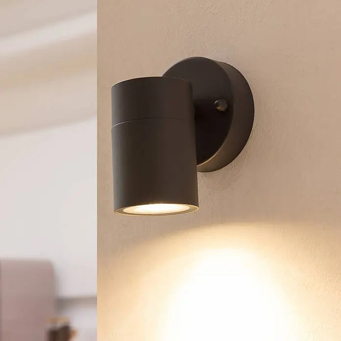 Prios Außenwandleuchte Tulimar - schwarz, schwarz, IP44 - Schwarze zylindrische Wandleuchte strahlt warmes Licht nach unten auf eine texturierte Wand.