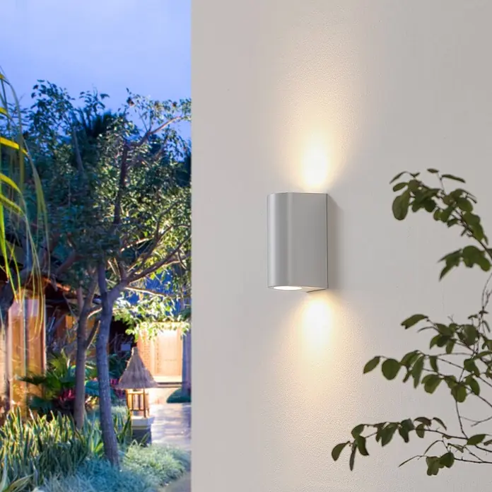 Prios Außenwandleuchte Irfan - weiß / opal, IP44 - Silberne Outdoor Up-Down Wandleuchte an einer strukturierten Wand, die warmes Licht nach oben und unten in einem Garten abgibt.