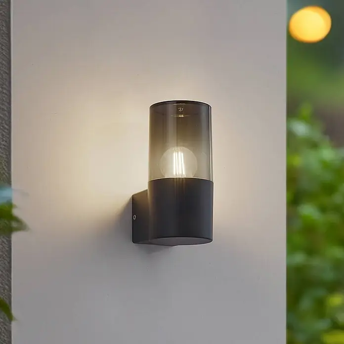 Lindby Außenwandleuchte Heulo - 10,8 x 8 x 16,7 cm, schwarz, schwarz, IP44 - Dunkelgraue Außenwandleuchte mit Rauchglaszylinder und leuchtender Filamentlampe, an einer Wand montiert.