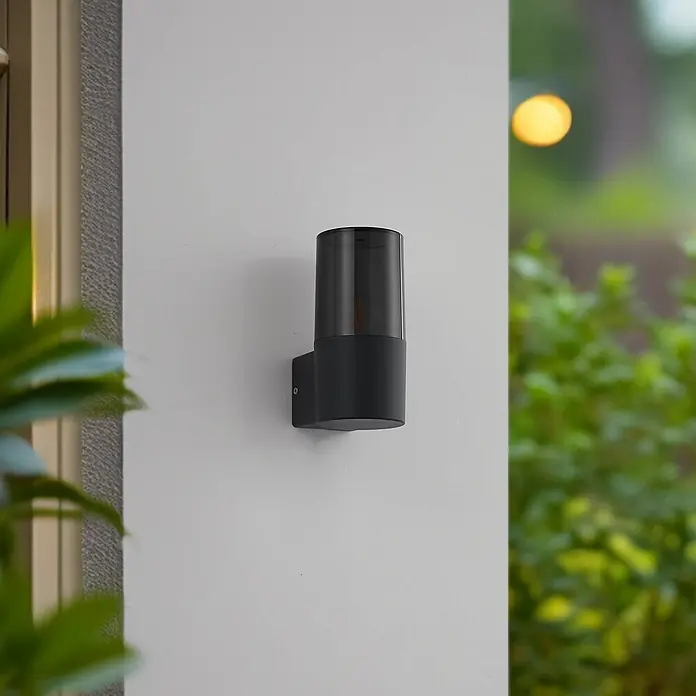 Lindby Außenwandleuchte Heulo - 10,8 x 8 x 16,7 cm, schwarz, schwarz, IP44 - Dunkelgraue Außenwandleuchte mit lichtdurchlässiger Oberseite, an einer hellen Wand montiert.