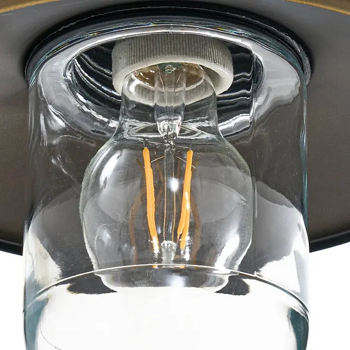 Lindby Außenwandleuchte Eloretta - messing / gold, messing / gold, IP44 - Nahaufnahme einer Leuchte mit klarem Glasschirm, weißer Fassung und LED-Filamentlampe, auf dunklem Metallsockel.