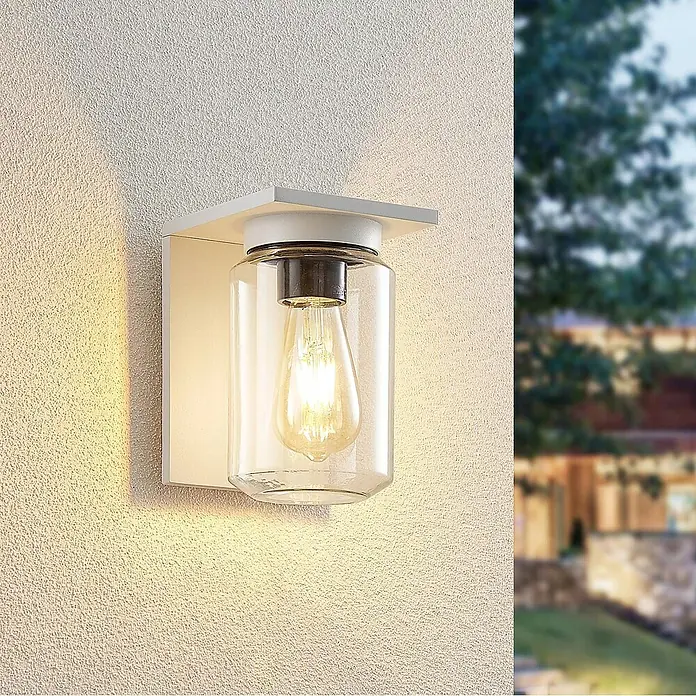 Lucande Außenwandleuchte Semka - weiß / opal, klar / transparent, IP54 - Moderne weiße Wandleuchte mit klarem Glaszylinder und sichtbarer Glühbirne, montiert an einer strukturierten Außenwand.