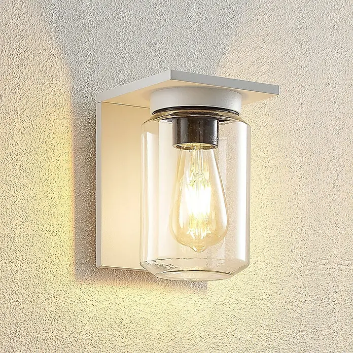 Lucande Außenwandleuchte Semka - weiß / opal, klar / transparent, IP54 - Weiße Metall-Wandleuchte mit klarem Glaszylinder und beleuchteter Edison-LED-Birne, die warmes Licht auf eine texturierte Wand wirft.