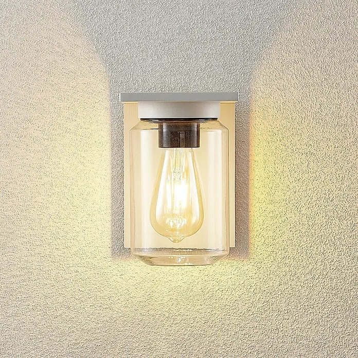 Lucande Außenwandleuchte Semka - weiß / opal, klar / transparent, IP54 - Wandleuchte mit klarem Glasgefäß und sichtbarer Edison-Birne, montiert an strukturierter Wand, wirft warmes Licht.