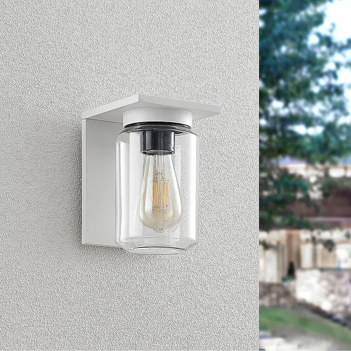 Lucande Außenwandleuchte Semka - weiß / opal, klar / transparent, IP54 - Weiße Außenwandleuchte mit klarem Glaszylinder und Edison-LED-Lampe, an einer strukturierten weißen Wand montiert.