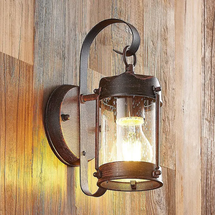 Lindby Außenwandleuchte Farly - 17,3 x 9,2 x 26 cm, braun / rost, IP23 - Rustikale Wandlaterne aus braunem Metall mit Samenglas, spendet warmes Licht an Holzwand.