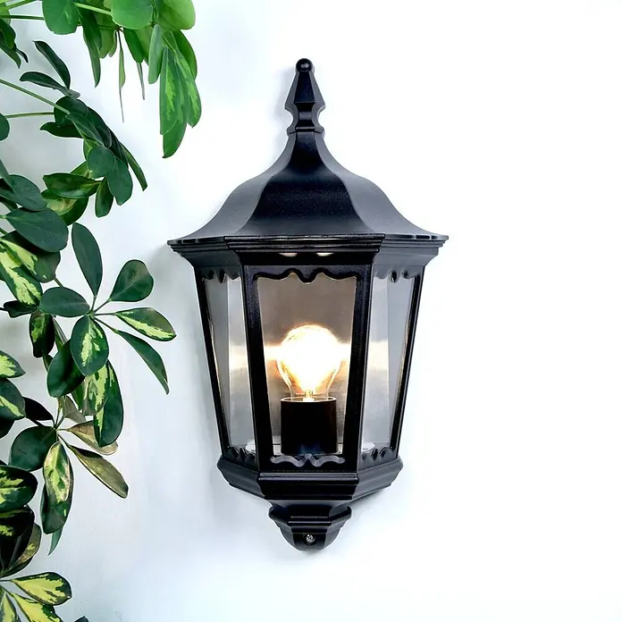 Lindby Wandleuchte aussen in schwarz, transparent, Aussenlampe Wand -  - Schwarze Außenwandlaterne mit leuchtender Glühbirne, umgeben von grünem Blattwerk.