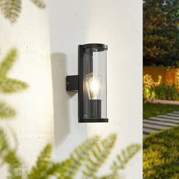 Lucande Außenwandleuchte Zanta - schwarz, alu / grau / zink, IP65 - Outdoor-Wandleuchte mit schwarzem Rahmen und klarem Glaszylinder an einer strukturierten Wand im Garten.